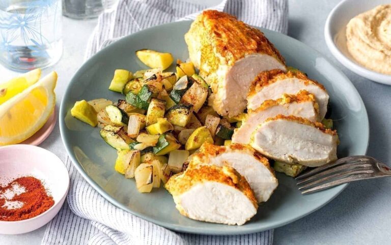 A plate of food with sliced, baked chicken breast, accompanied by diced and grilled zucchini and yellow squash. There is a lemon wedge, a bowl of hummus, and a small bowl of paprika on the side. A fork rests on the edge of the plate. MyFitnessPal Blog